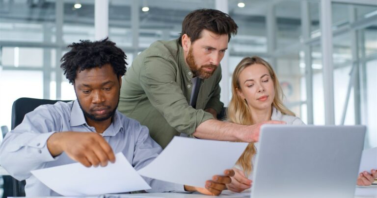 Three professionals reviewing paper documents beside a laptop in a bright office.